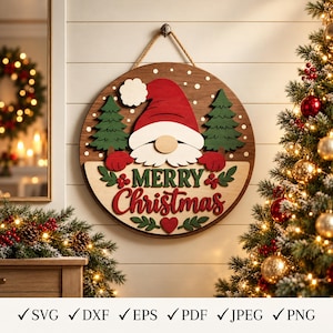 Könnte beinhalten: Ein rundes Wand-Schild aus Holz mit einem Wichtel, der eine rote Mütze, einen weißen Bart und rote Fäustlinge trägt, flankiert von zwei grünen Tannen. Das Schild zeigt den Text Merry Christmas in Rot und Grün, verziert mit weißen Schneeflocken.