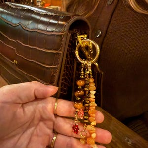 May include: A dark brown crocodile-textured leather handbag featuring a gold-toned metal ring attachment. Dangling from the ring are three beaded strands with tiger eye stones, orange chips, and small red leaf-shaped metal charms.