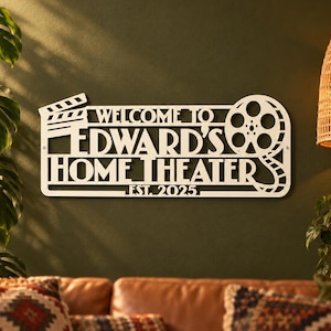 May include: A white wall-mounted sign featuring a film clapperboard icon on the left and a film reel on the right. The text reads "WELCOME TO EDWARD'S HOME THEATER EST. 2025" in a bold, cut-out font style, creating a decorative cinema-themed display.