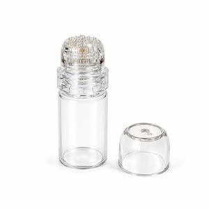 Op de afbeelding: Een doorzichtige, cilindrische glazen fles met een speciale applicator-kop met talloze kleine, scherpe metalen naaldjes aan de bovenkant. De fles bevat een aparte, gladde, transparante, koepelvormige dop die ernaast is geplaatst.