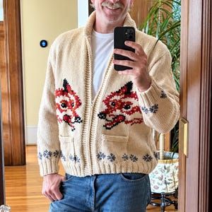 May include: A cream-colored knit zip-up cardigan featuring a repeating fox face pattern in red, black, and white across the chest. The garment includes a ribbed collar, cuffs, and hem, with two front pockets and a grey snowflake border at the base.