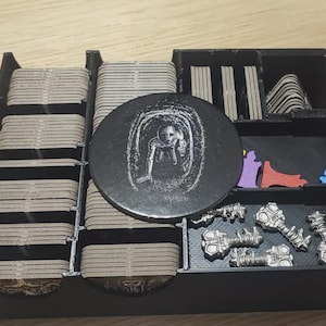 以下が含まれることがあります： 黒いプラスチック製のボードゲーム用オーガナイザー。長方形のカードボードタイル、トンネル内の人物が白く描かれた黒い円形トークン、銀色の金属製キー4本、紫・赤・青の小さな形状のパーツが収納されています。