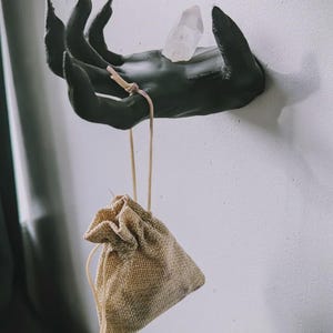 May include: A matte black wall-mounted hand sculpture with long, pointed fingers. A clear, translucent quartz crystal rests on the palm. A small, tan-colored burlap drawstring pouch hangs from the middle finger by a thin, light-colored cord.