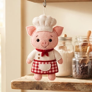 Close-up of a pink felt Baker Pig showing the chef hat, embroidered eyebrows and smile, rose-colored nose, safety eyes, red neckerchief, and checkered apron.