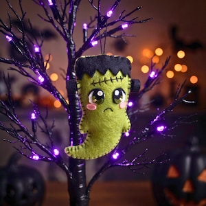 Green felt Frankenstein ghost Halloween ornament hanging from a dark mantel with garland, candles and purple lights.
