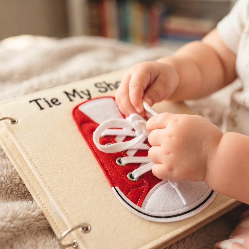 Close-up of a toddler's hands practicing shoe tying on a handmade felt quiet book page featuring a red sneaker and white laces.