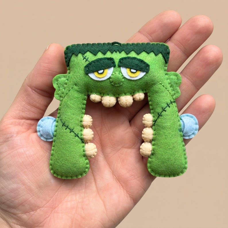 Hand holding a green felt Frankenstein monster ornament to show its 5-inch size against a beige background.