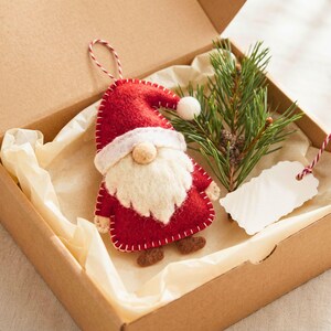 Felt Santa gnome Christmas ornament nestled in a kraft gift box with cream tissue paper, a pine sprig and a blank gift tag.