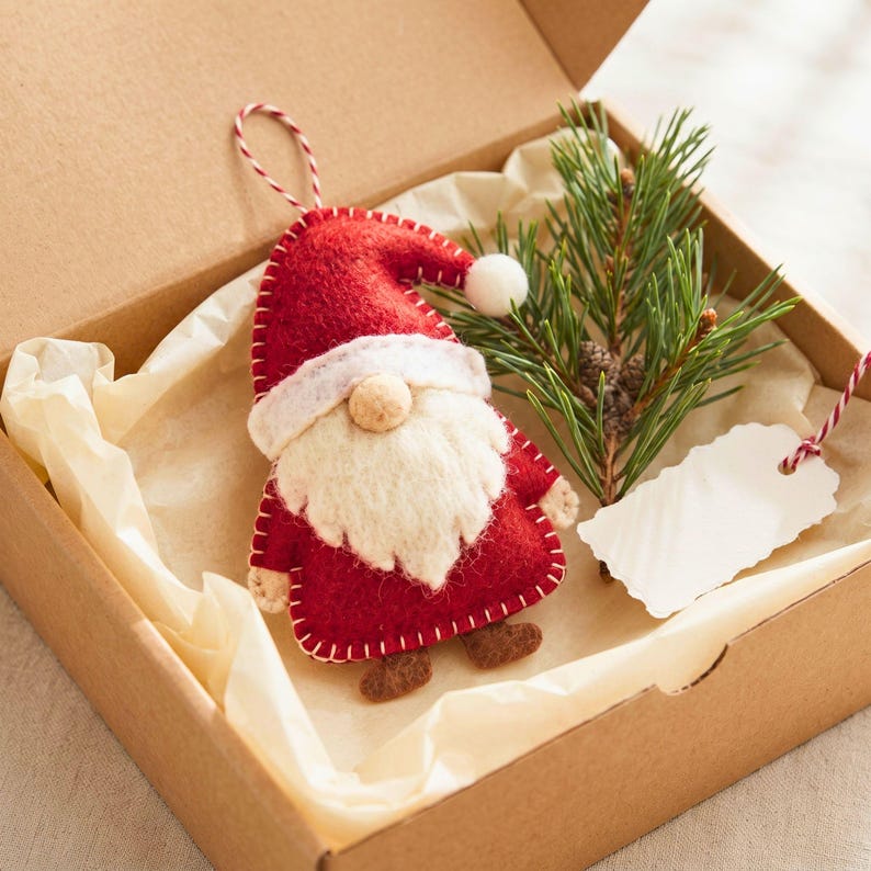 Felt Santa gnome Christmas ornament nestled in a kraft gift box with cream tissue paper, a pine sprig and a blank gift tag.