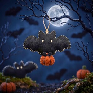 Black felt bat ornament holding a tiny pumpkin hanging from a bare branch under a full moon