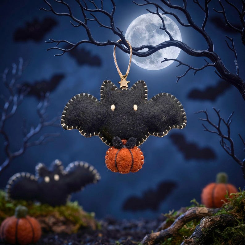 Black felt bat ornament holding a tiny pumpkin hanging from a bare branch under a full moon