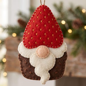 Handmade felt gnome ornament shown from another view with a red strawberry hat, cream beard, brown body, and decorative seed stitching.