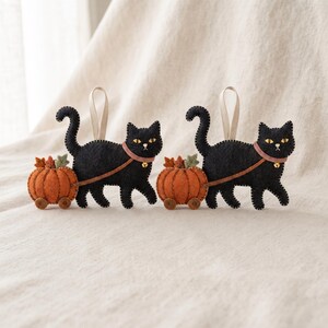Two handmade felt black cat ornaments displayed together on textured neutral linen fabric.
