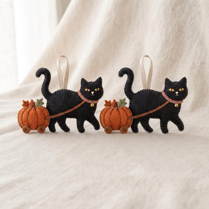 Two handmade felt black cat ornaments displayed together on textured neutral linen fabric.