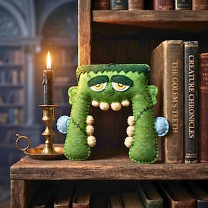Green felt Frankenstein monster ornament on a bookshelf between horror books beside a lit black candle.