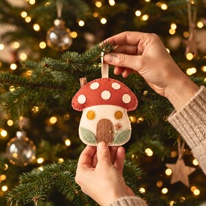 Handmade felt mushroom house hanging ornament displayed on a rustic Christmas tree branch.