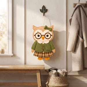 Finished librarian owl felt ornament in a cozy decorative setting, showing the tan owl body, cream face, dark glasses, green clothing, and plaid skirt.