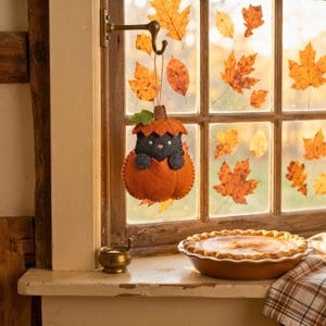 Felt cat in pumpkin ornament hanging in a farmhouse kitchen window beside a pumpkin pie