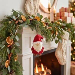 Felt Santa gnome ornament hanging from a pine and eucalyptus garland on a white fireplace mantel, with stockings and candles blurred behind it.