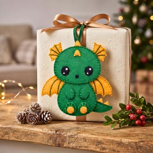 Finished green and golden-yellow felt dragon hanging from a loop as a decorative ornament, showing the dragon’s wings, tail, feet and embroidered details.