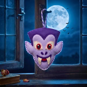 Lavender felt vampire ornament with fangs hanging in a window with a full moon and bat silhouettes behind it.
