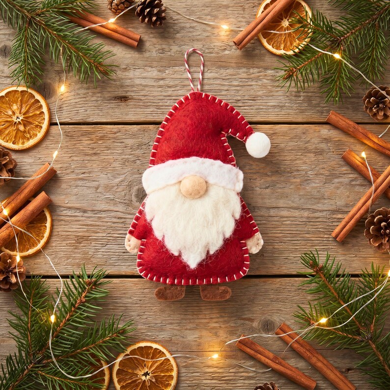 Felt Santa gnome ornament in a rustic Christmas flat lay with pine sprigs, cinnamon sticks, dried orange slices and pinecones on a wooden table.