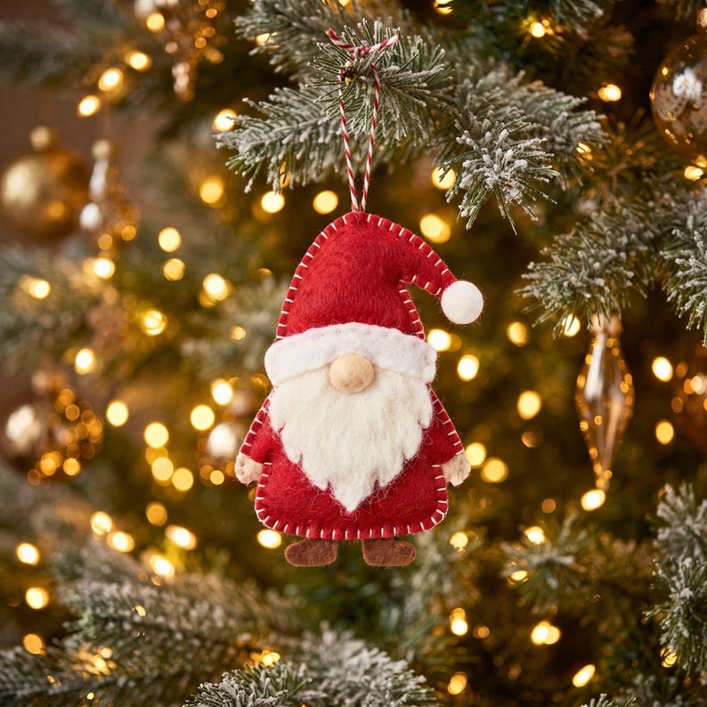 Felt Santa gnome ornament hanging from a frosted pine branch on a Christmas tree, with warm golden fairy lights softly blurred in the background.