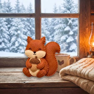 A handcrafted felt squirrel holding a brown acorn sits on a wooden windowsill beside a mug and a knitted blanket against a snowy window background.