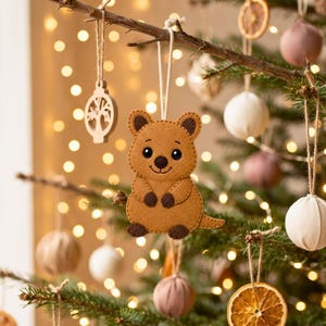Cute handmade tan felt quokka ornament with rounded ears, dark brown details, smiling face, arms, feet, and curved tail on a light background.