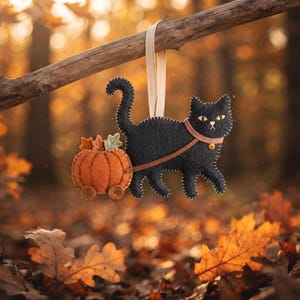 Handmade charcoal black felt cat ornament pulling a tiny pumpkin cart hanging from a rustic branch with autumn decor.