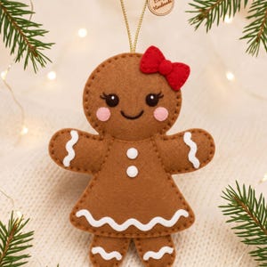 Close-up of a felt gingerbread girl face with black eyes, eyelashes, smiling mouth, pink cheeks, red bow and white decorative details.