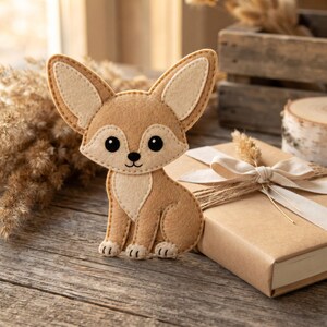 Cute fennec fox felt ornament showing the sandy tan body, large cream-lined ears, cream muzzle, embroidered smile, and hanging loop.