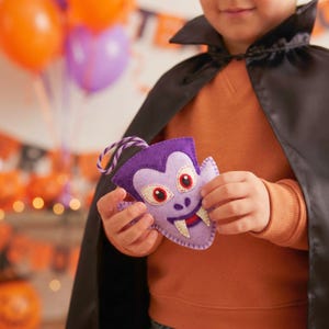 Child in a vampire cape holding a lavender felt vampire ornament at a Halloween party.