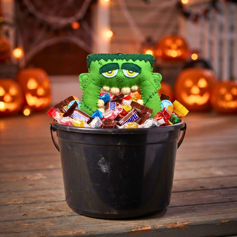 Green felt Frankenstein monster ornament peeking out of a candy-filled trick-or-treat bucket on a porch at dusk.