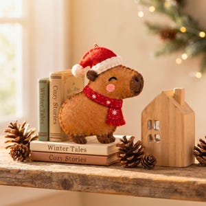 Felt capybara ornament styled on a wooden shelf next to holiday pinecones and miniature cabin decor.