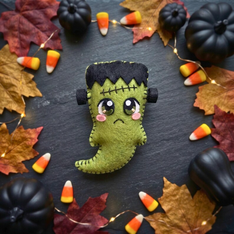 Overhead view of a green felt Frankenstein ghost ornament styled with candy corn, mini pumpkins and fairy lights.