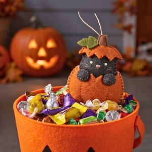 Felt cat in pumpkin ornament perched on a trick-or-treat bucket full of candy