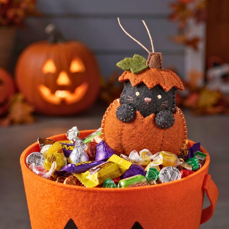 Felt cat in pumpkin ornament perched on a trick-or-treat bucket full of candy