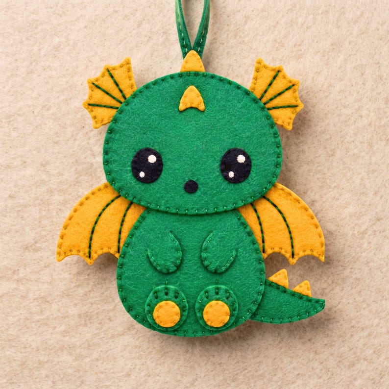 Close-up of a kawaii felt dragon face with black eyes and nose, white eye highlights, green felt head, decorative crest and golden-yellow wing details.
