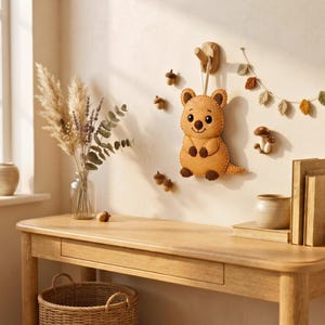 A brown felt quokka toy hangs on a light wall above a wooden desk styled with dried grasses, small felt acorns, and a leaf garland.