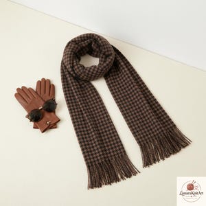 May include: A brown and black houndstooth patterned scarf with fringed ends, paired with a pair of tan leather gloves and dark-tinted sunglasses. The scarf features a repeating check design and is displayed alongside the accessories.