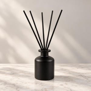 Puede incluir: Un difusor de varillas de color negro mate con una base cilíndrica y un cuello estrecho. Cinco varillas negras finas y rectas están insertadas en la abertura de la botella, extendiéndose hacia arriba en forma de abanico.