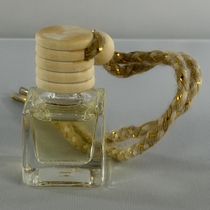 May include: A small, square, clear glass bottle filled with a light-colored liquid. The bottle features a cylindrical, light-colored wooden cap with horizontal grooves and a thick, braided beige rope cord interwoven with thin, metallic gold threads.