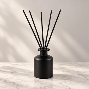 May include: A matte black reed diffuser bottle with a cylindrical base and a narrow neck. Five thin, straight black diffuser reeds are inserted into the opening of the bottle, extending upwards in a fan-like arrangement.