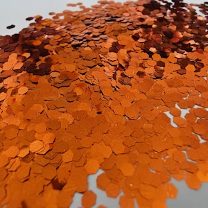 May include: A collection of small, metallic orange hexagonal glitter pieces scattered across a surface. The individual flakes have a reflective, shiny finish and vary slightly in their shade of orange, creating a dense, textured pile of craft material.