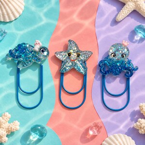 May include: Three blue metal paper clips topped with resin sea creatures. One features a blue glitter sea turtle with a small white figure, one a blue glitter starfish with a smiling face, and one a blue glitter octopus with a small pink crown.