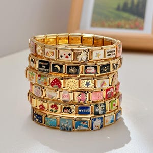 May include: A stack of gold-tone modular link bracelets featuring various colorful enamel charms. The charms display diverse designs including flowers, fruits, celestial symbols, spiders, and text such as DREAM, I LOVE MY BOYFRIEND, and I LOVE JESUS.