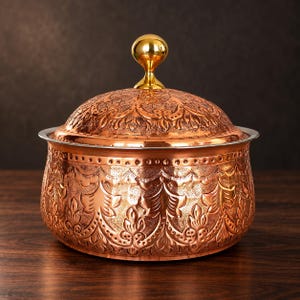 Peut inclure: Un bol de service rond en cuivre avec un couvercle assorti, présentant des motifs floraux et végétaux en relief sur toute la surface. Le couvercle est surmonté d'un bouton métallique doré, sphérique et poli, avec un bord large et lisse.