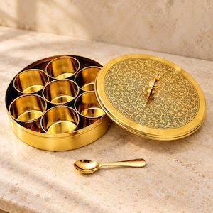 Op de afbeelding: Een ronde goudkleurige metalen kruidendoos met zeven kleine uitneembare ronde bakjes aan de binnenkant. Het deksel heeft een ingewikkeld zwart bloemmotief en een centrale knop. Een klein bijpassend goudkleurig metalen lepeltje ligt ervoor.