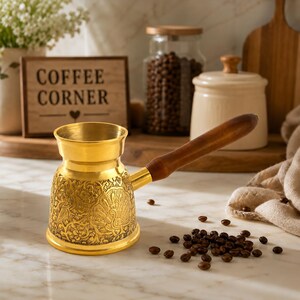 Könnte beinhalten: Eine goldfarbene Kaffeekanne aus Metall mit einem detaillierten Pfauen- und Blumenmuster auf dem Korpus, ergänzt durch einen langen, glatten, dunkelbraunen Holzgriff an der Seite, konzipiert für die Kaffeezubereitung auf dem Herd.